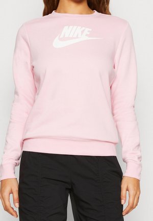 Osoba ubrana w jasnoróżową bluzę Nike z długim rękawem i czarne spodnie, stoi z ramionami luźno opuszczonymi wzdłuż ciała.