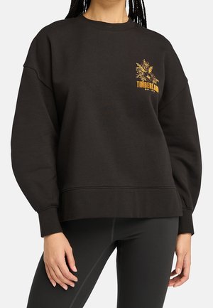 Kobieta w czarnej bluzie Timberland z żółtym kwiatowym logo na piersi, w połączeniu z czarnymi legginsami, stojąca na jednolitym tle.