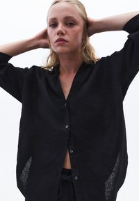 Czarna lniana koszula z długimi rękawami, z zapięciem na guziki, otwartym kołnierzykiem i bocznymi rozcięciami. Posiada kieszeń na piersi oraz teksturowany materiał.