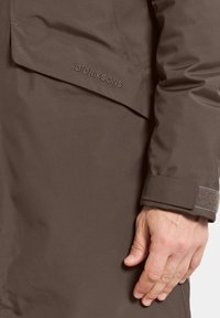 Ruskea vedenpitävä takki, jossa on sileä pinta, hihansuu tarrakiinnityksellä ja hienovarainen rintamerkki kirjailtuna.