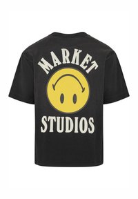 Melna T-kreka ar lielu dzeltenu smaidošu seju grafiku, ar uzrakstu "MARKET" augšpusē un "STUDIOS" apakšpusē treknajā baltajā tekstā. Īsas piedurknes, brīvs griezums.