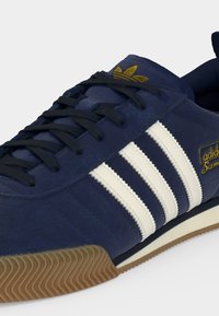 Baskets Adidas Samba bleu marine avec bandes blanches, lacets noirs, logo Adidas doré sur la languette, texte doré "Samba" sur le côté et semelle en gomme.