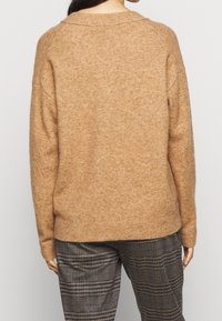 Brun stickad tröja med lös passform, bred krage och ribbad nederkant; mjuk textur och subtil melerat mönster. Matchas med rutiga byxor.