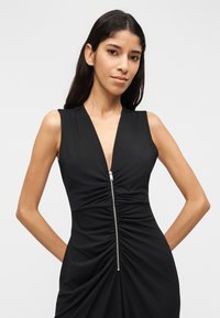 Γυναίκα με μακριά σκουρόχρωμα μαλλιά που φοράει αμάνικο μαύρο φόρεμα με φερμουάρ μπροστά και ρυτιδωμένη λεπτομέρεια, στέκεται μπροστά σε απλό φωτεινό φόντο.