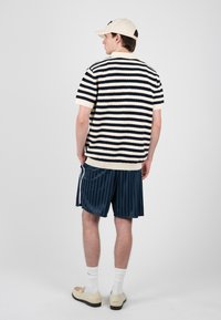 Homme portant une casquette beige, un pull à manches courtes rayé bleu marine et blanc, un short rayé bleu marine, des chaussettes blanches et des mocassins beiges, vu de dos.