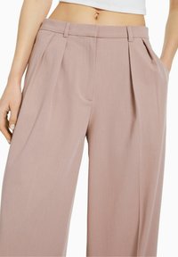 Ljust rosa vida byxor med veck, som har en slät yta och en traditionell midja. Fickor är synliga på båda sidor.