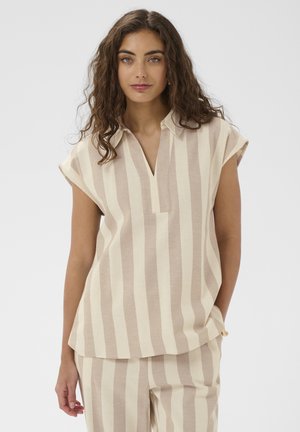 Mujer con cabello rizado que lleva una camisa de manga corta de rayas verticales beige y crema y pantalones a juego, posando frente a un fondo liso.