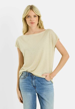 Joven mujer con cabello rubio, vistiendo una camiseta beige de manga corta y vaqueros azules, posando con una mano en el bolsillo frente a un fondo blanco.