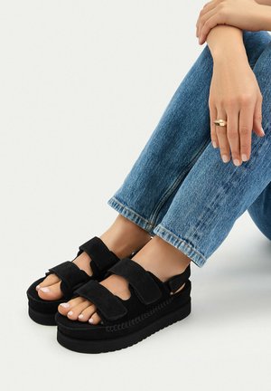 Stopy w czarnych sandałach na platformie z dwoma paskami na rzep, ubranych w niebieskie dżinsy, ręce spoczywające na skrzyżowanych kolanach, złoty pierścionek na palcu.