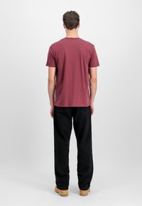 Karmīna īsām piedurknēm T-krekls ar taisnu griezumu, kopā ar melniem biksēm un brūniem apaviem, skatīts no aizmugures uz neitrāla fona.