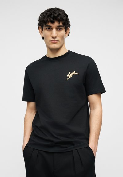 Νέος άνδρας με κοντά σγουρά μαλλιά που φοράει μαύρο μπλουζάκι με ένα μικρό μπεζ λογότυπο λαγού που πηδάει στο πάνω αριστερό μέρος του στήθους, με τα χέρια στις τσέπες.