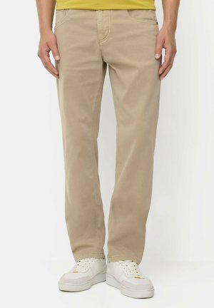 Unterkörper einer Person, die beige Hose, weiße Sneaker und ein gelbes Hemd trägt und vor einem schlichten weißen Hintergrund steht.