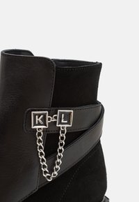 Černé kotníkové boty kombinující kůži a semiš. Výrazný stříbrný řetěz spojuje čtvercové ozdoby s písmeny "K" a "L".