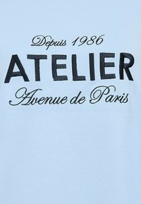 Light blue cotton T-shirt featuring black embroidered text: "ATELIER," "Depuis 1986," and "Avenue de Paris" in decorative fonts.
