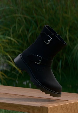 Stivaletto nero alla caviglia con fibbie, realizzato in materiale testurizzato, con suola robusta e design essenziale, ambientato su uno sfondo di verde naturale.