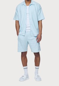 Chemise bleu clair à manches courtes et short assorti en tissu texturé, associés à des sandales blanches à enfiler et des chaussettes blanches.