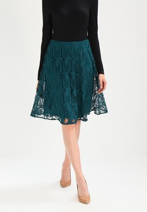 Femme portant une jupe en dentelle bleu sarcelle longueur genou, un haut à manches longues noir, et des talons hauts beige, debout avec une jambe croisée devant.
