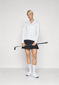 Fehér, cipzáros, hosszú ujjú felső, fekete szoknya és fehér sportcipő. Golfütőt tart a kezében. Sima anyag, egyszerű dizájn, térd fölé érő szoknya.