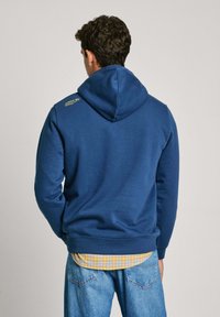 Kapucna majica v mornarsko modri barvi, izdelana iz mehke tkanine. Ima rumen logotip na rami in rebrovan rob, nameščena čez karo srajco.