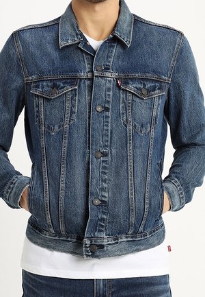 Mężczyzna noszący niebieską dżinsową kurtkę z zapięciem na guziki, klapami na kieszeniach na piersi oraz rękoma w bocznych kieszeniach, na białej koszuli.