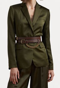 Olivově zelený blazer s klopou, hnědý kožený opasek s detailem ve zlatém řetězu a dvěma předními chlopeňovými kapsami. Hladký povrch látky.