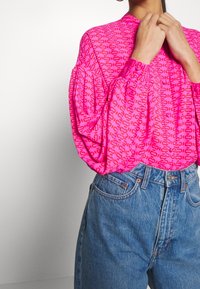 Różowa bluzka w wzory z bufkami, zestawiona z jeansami typu high-waisted w niebieskim kolorze. Bluzka wyróżnia się powtarzającym się wzorem logo.