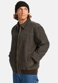 Giacca in corduroy marrone con zip, dotata di colletto a risvolto, tasche frontali e polsini a costine. Indossata sopra un maglione scuro e jeans blu.