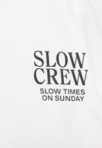 Črni napis na belem blagu pravi "SLOW CREW" z manjšim napisom spodaj, ki pravi "SLOW TIMES ON SUNDAY."