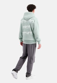 Mintgrüner Hoodie mit weißem Text "STARTED FROM NOTHING" auf dem Rücken, kombiniert mit grauen Cargo-Hosen und weißen Sneakers. Weicher Stoff.