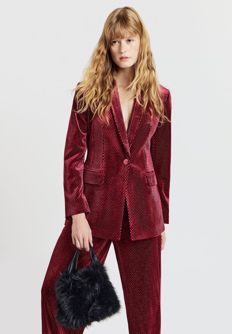 Abito in velluto cremisi con blazer aderente, rever a intaglio e due tasche frontali, abbinato a pantaloni coordinati e una piccola borsa nera in ecopelliccia.