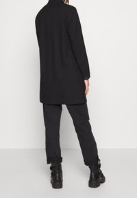 Abrigo largo negro de corte recto, con cuello con solapa y mangas remangadas. Combinado con pantalones oscuros de corte holgado y botas negras gruesas.