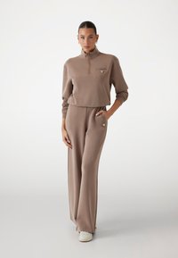 Lysebrune to-dels outfit bestående af en lynlås-langærmet top og brede bukser, lavet af blødt materiale med diskrete logo detaljer.