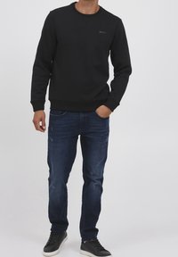 Felpa nera con collo a giro e polsini a coste, abbinata a jeans scuri invecchiati e sneakers nere, in piedi contro uno sfondo semplice.