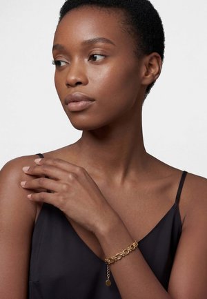 Lyhyttukkainen nainen, jolla on musta hihaton toppi ja kultainen ketjurannekoru, katsoo vasemmalleen käden levätessä olkapäällä.