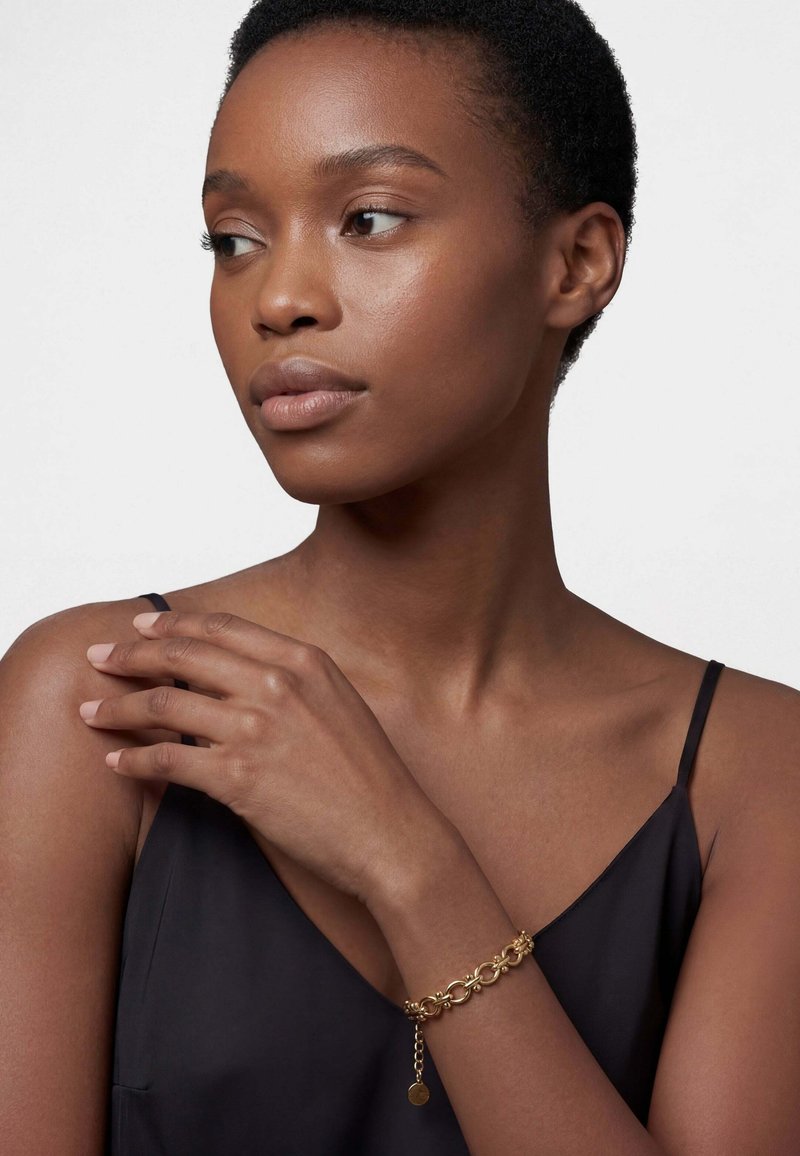 Rövid, göndör hajú nő fekete ujjatlan felsőben és arany lánckarkötőben, balra nézve, kezét a vállán pihentetve.