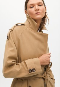Beige trenchcoat med hög krage, knäppta ärmslut och bälte i midjan. Tillverkad av ett mjukt tyg med subtil lyster och strukturerad design.