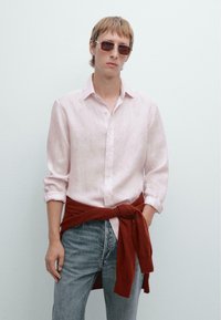 Lichtroze linnen shirt met opgerolde mouwen, gecombineerd met donkerblauwe denim jeans en een rode gebreide trui om de taille geknoopt, aangevuld met een zonnebril.