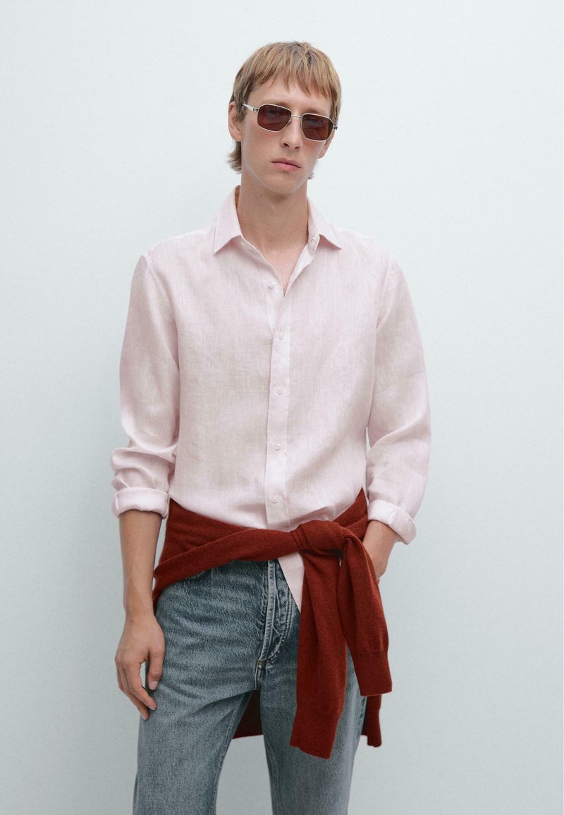 Lichtroze linnen shirt met opgerolde mouwen, gecombineerd met donkerblauwe denim jeans en een rode gebreide trui om de taille geknoopt, aangevuld met een zonnebril.