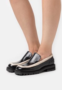 Chaussures noires en cuir à enfiler avec un accent beige le long du bord, dotées d'une semelle épaisse et d'un design à bout arrondi, présentant une texture lisse.