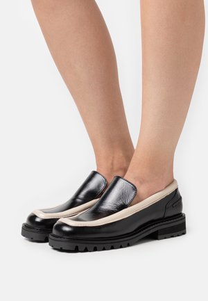 Chaussures noires en cuir à enfiler avec un accent beige le long du bord, dotées d'une semelle épaisse et d'un design à bout arrondi, présentant une texture lisse.