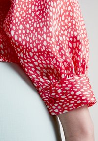 Blusa rossa con motivo a pois bianchi, caratterizzata da un polsino con bottoni e dettagli arricciati, realizzata in tessuto leggero e liscio.