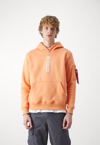 Sudadera con capucha en naranja pastel, hecha de un tejido suave. Presenta un parche logo central y un bolsillo. Incluye una etiqueta en la manga lateral con acento rojo.