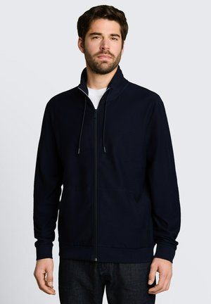 Man met kort donker haar en baard, draagt een zwart jack met rits over een wit overhemd, staand tegen een grijze achtergrond.