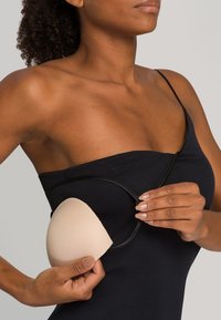 Coppa per reggiseno adesiva beige con texture liscia, circondata da una sottile fascia nera, tenuta da una persona che indossa una maglietta senza maniche nera.