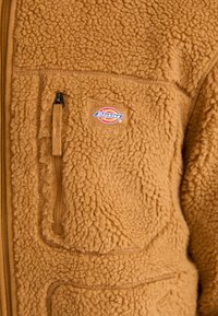 Brown fleece jacket with a textured, fluffy surface, featuring a chest pocket and a visible Dickies logo patch. Zipper closure.