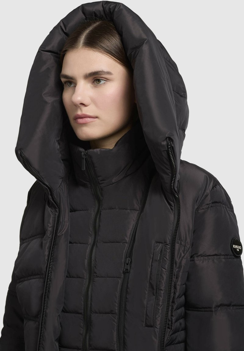 Doudoune noire avec une grande capuche surdimensionnée, texture matelassée, fermetures éclair à l'avant et une poche latérale avec un logo.
