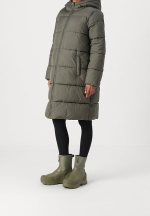 Persoon draagt een olijfgroene, knielange donsjas met capuchon, zwarte legging en bijpassende olijfgroene halfhoge laarzen met ritssluitingen aan de zijkant.