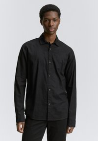 Zwarte lange mouwen shirt in een gestructureerde stof, met een knoopsluiting aan de voorkant en een relaxte pasvorm. De kraag is standaard en de manchetten zijn met knopen.