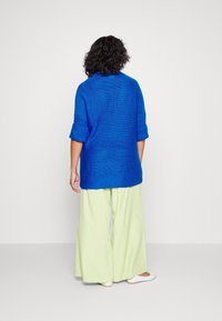 Överdimensionerad blå stickad tröja med rullade ärmar, kombinerad med ljusgröna vida byxor. Slät textur, avslappnad passform, synlig sömnad.