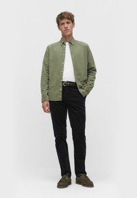 Vaaleanvihreä nappikauluspaita valkoisen t-paidan päällä, yhdistettynä tummiin housuihin ja ruskeisiin kenkiin, seisoo neutraalilla taustalla.